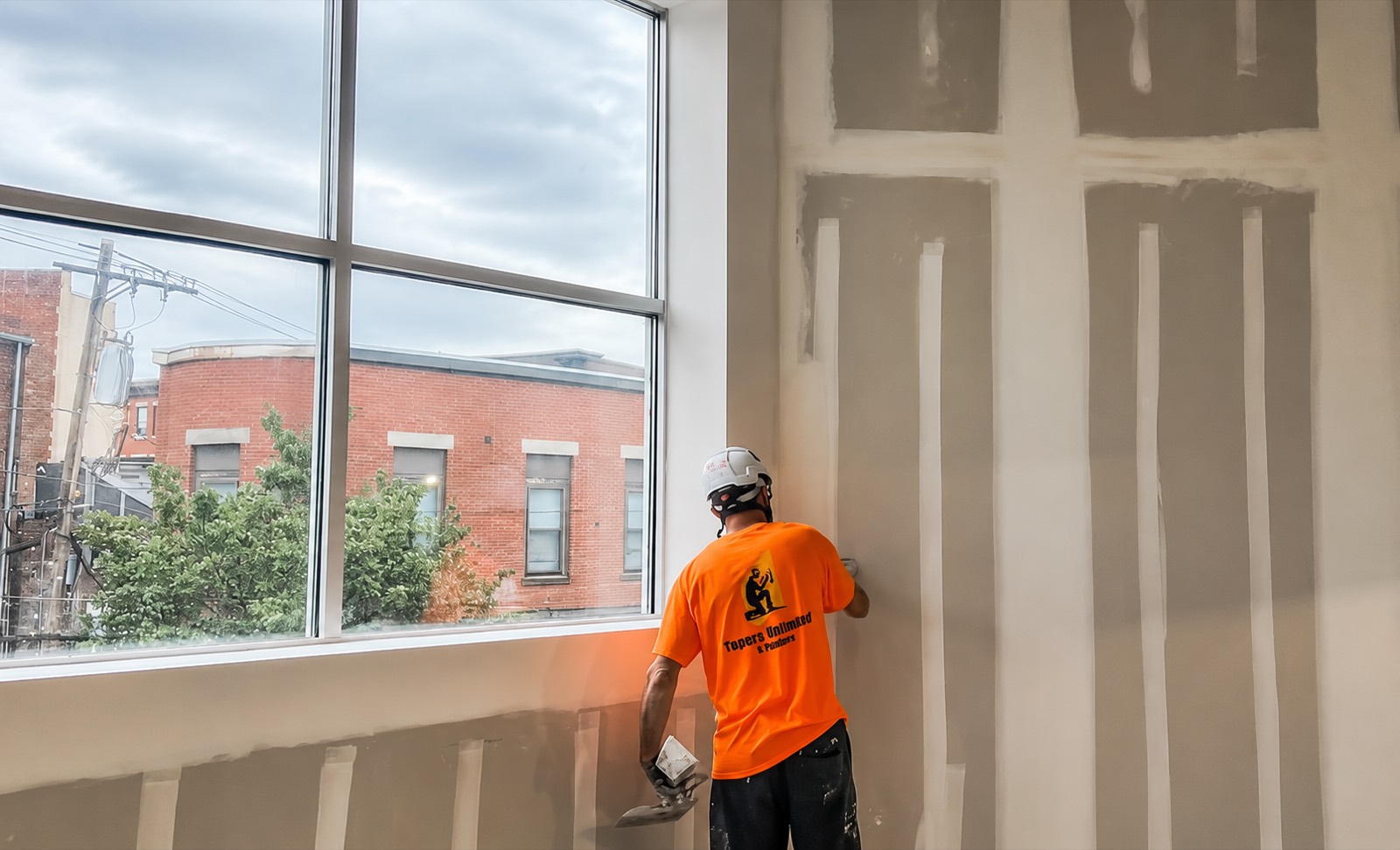 A Tapers Unlimited & Painters crew member in a branded orange shirt and hard hat finishing drywall seams on a tall commercial wall beside a window.