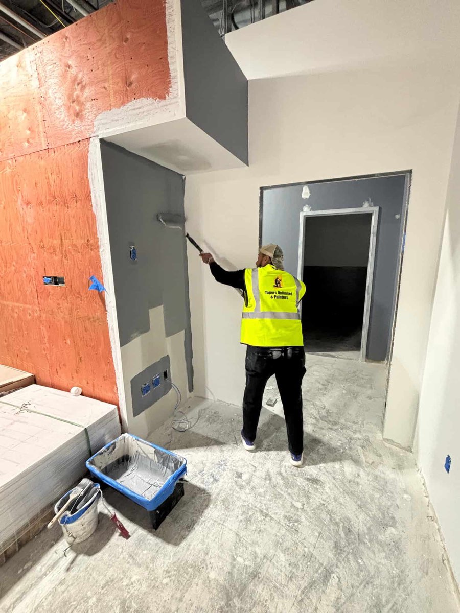 A Tapers Unlimited & Painters crew member in a branded hi-vis vest rolling grey paint onto a wall with an extension pole in a commercial fit-out, beside a stack of drywall board and a lined roller tray.