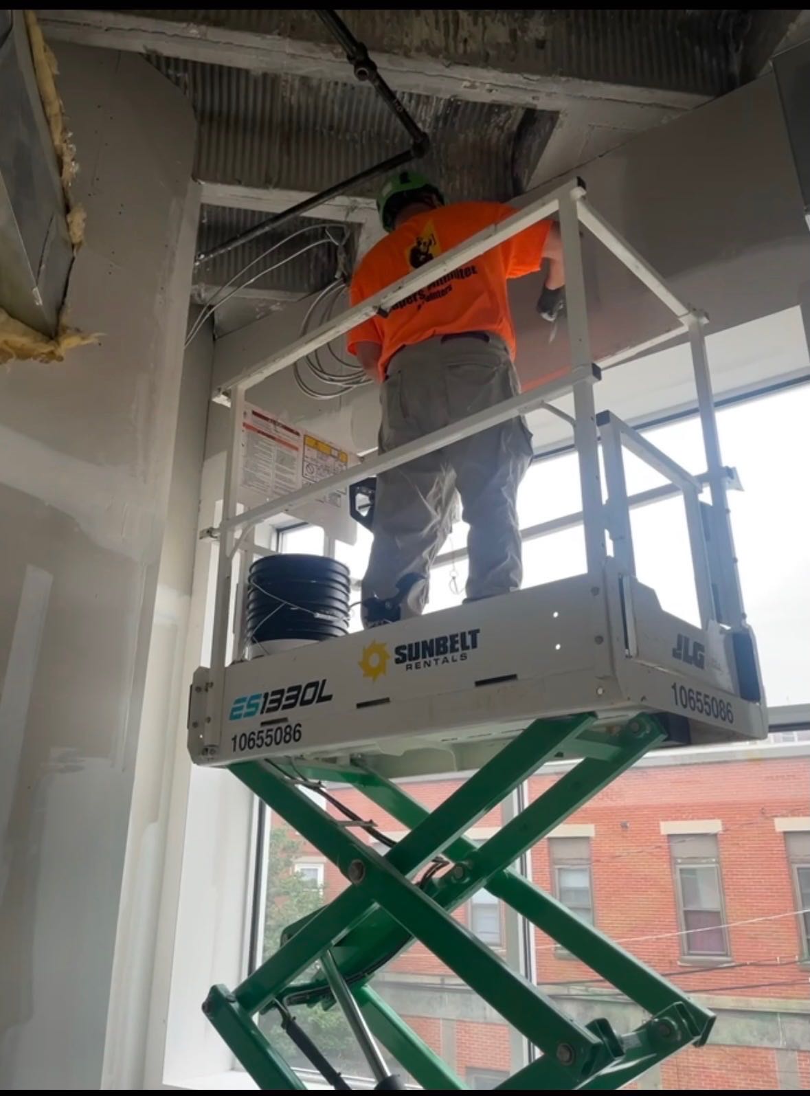 A Tapers Unlimited & Painters crew member in a branded orange shirt and green hard hat working from a scissor lift, taping the ceiling line of a commercial fit-out with a knife.