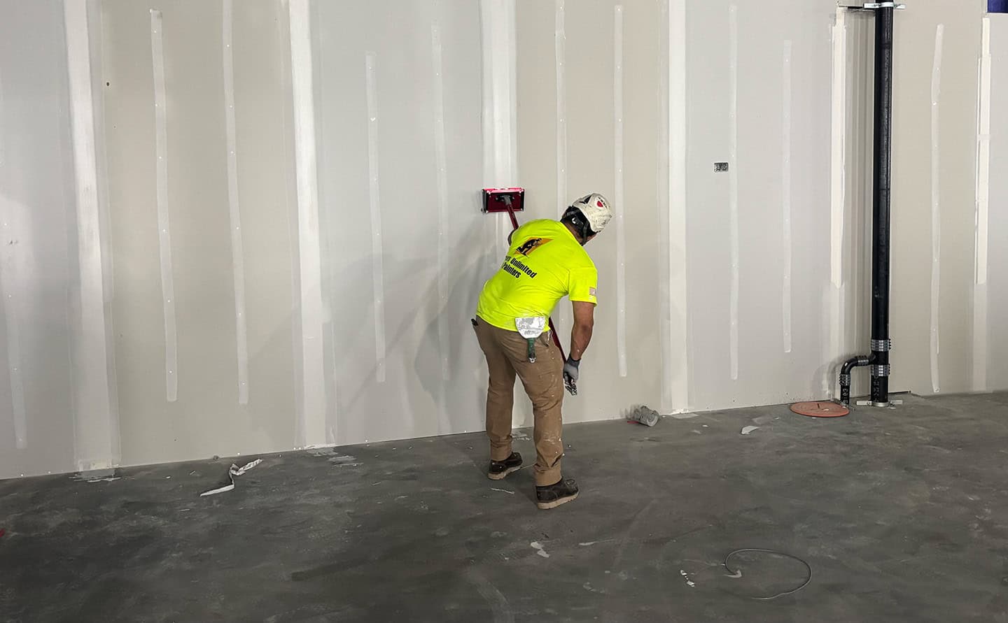 A Tapers Unlimited & Painters crew member in a branded hi-vis yellow shirt and hard hat running a flat box up a drywall seam, with freshly coated vertical seams across the finished wall.
